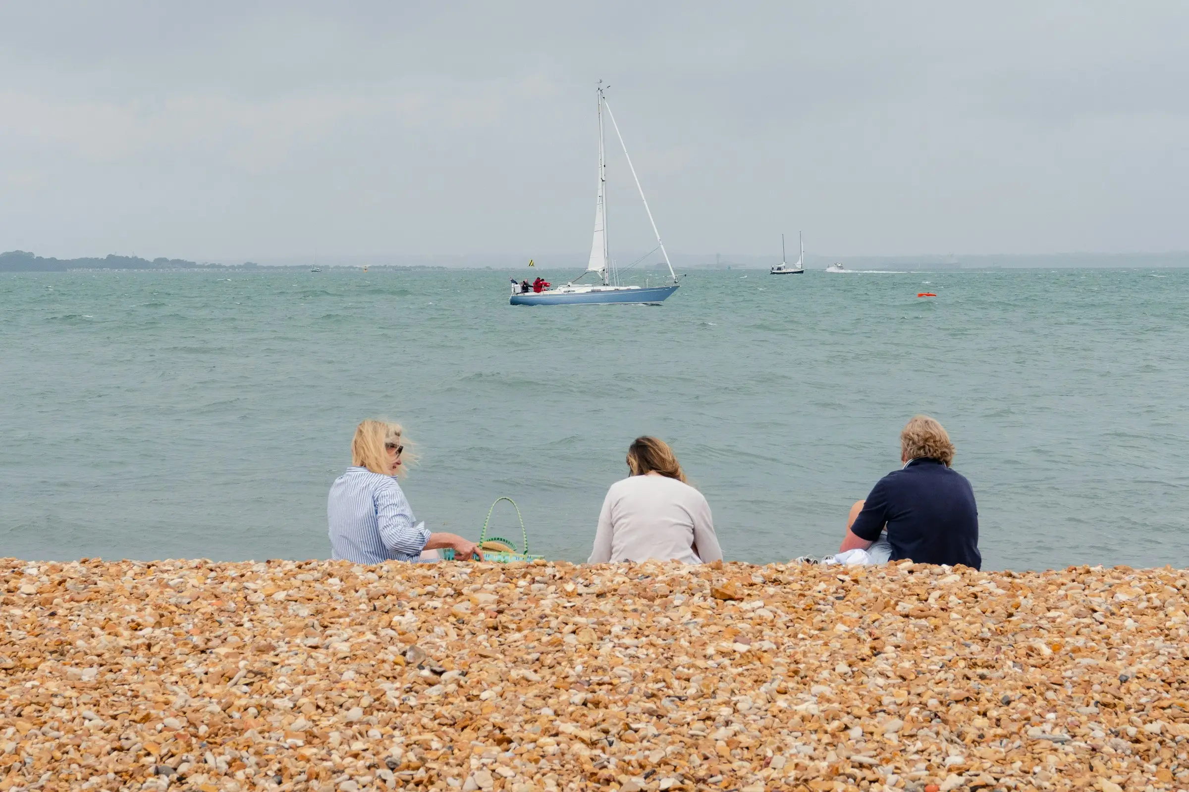 Three people sitting on the beach at Cowes, watching a yacht on the Solent